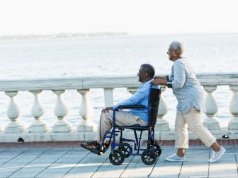 Woman pushes man in wheelchair by water.