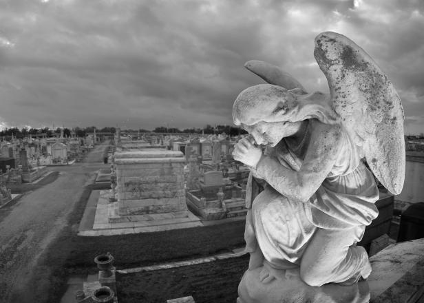 138186813 A praying statue kneels on top of a tomb in New Orleans under moody skies. High point of view to contain the cemetery landscape in the background.